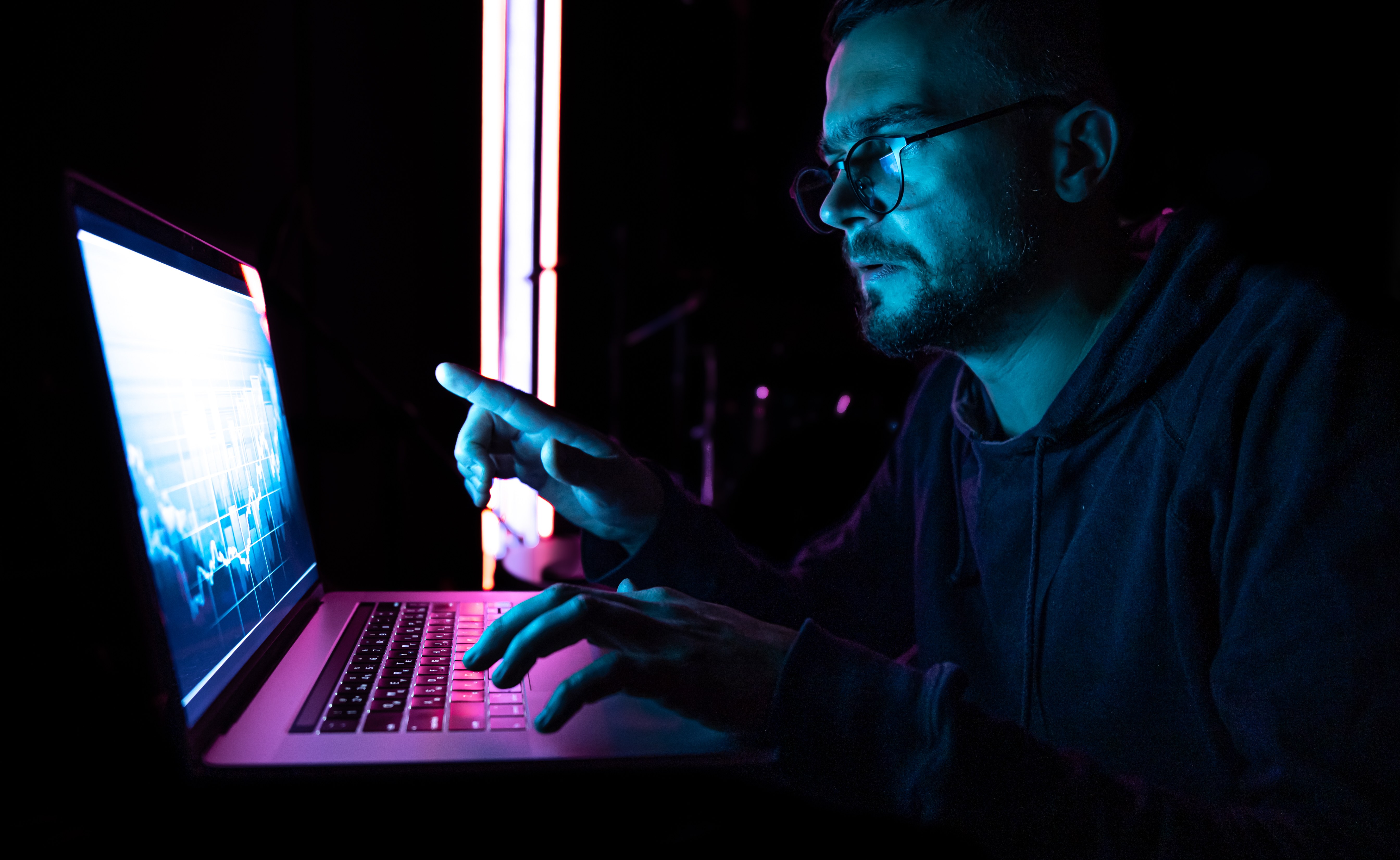 Homem digitando em um notebook com gráficos na tela, em ambiente escuro com luz neon